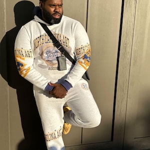 May include: A light gray hooded sweatshirt and matching sweatpants set featuring North Carolina A&T branding in blue and gold. The hoodie displays a central university emblem and text, while the pants feature matching text. Includes a black crossbody bag.