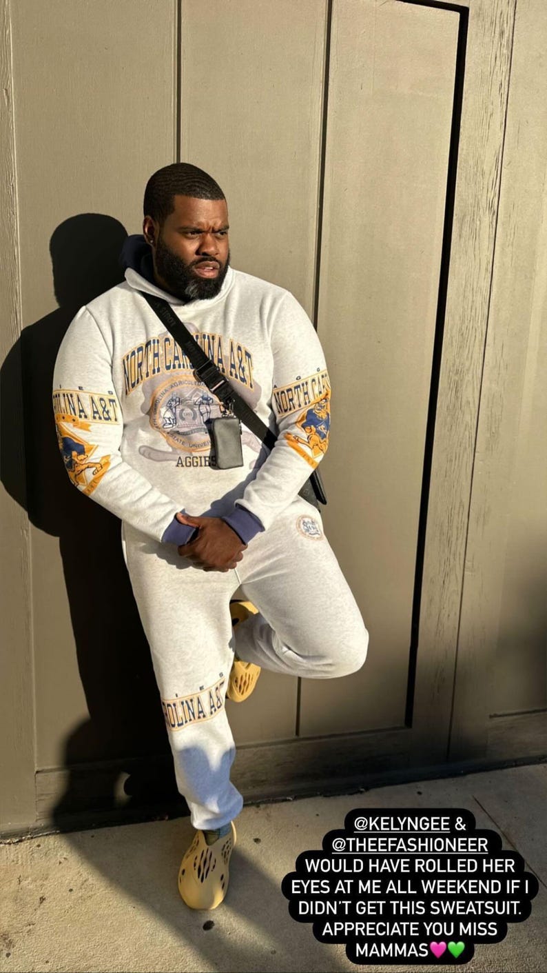 May include: A light gray hooded sweatshirt and matching sweatpants set featuring North Carolina A&T branding in blue and gold. The hoodie displays a central university emblem and text, while the pants feature matching text. Includes a black crossbody bag.