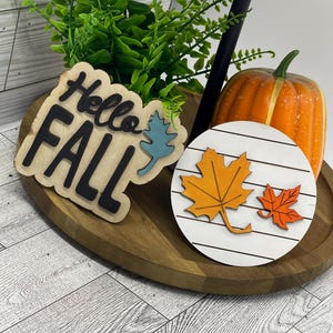 以下が含まれることがあります： 秋をテーマにした装飾が施された木製トレイ。 「Hello Fall」と書かれたサインと、2つの木製の秋の葉が付いた丸いサインが含まれています。 オレンジ色のカボチャと緑もあります。