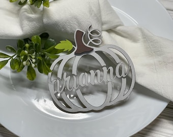 Puede incluir: Tarjeta de lugar de calabaza de madera blanca con el nombre "Arianne" escrito en negro. La calabaza está sobre un plato blanco con una servilleta blanca y follaje verde.