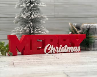Può includere: Un cartello rosso "MERRY" con testo bianco "Christmas", un piccolo albero di Natale glassato e un regalo confezionato. Il cartello è una decorazione festiva.
