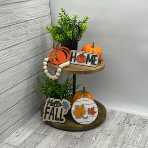 May include: A two-tiered wooden tiered tray with fall decor. The top tier has a wooden sign that says "HOME" with a pumpkin graphic. The bottom tier has a wooden sign that says "Hello FALL" with a leaf graphic. There are also two orange pumpkins and a string of white beads.