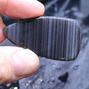 May include: A polished, black gemstone with parallel, light-colored striations. The stone has a rounded, rectangular shape with one pointed end. The surface is smooth and reflective, with a subtle sheen.