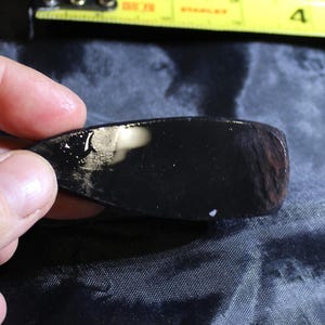 May include: A black, polished, smooth, and slightly curved piece of obsidian. The obsidian has a slight sheen and is held in a hand.