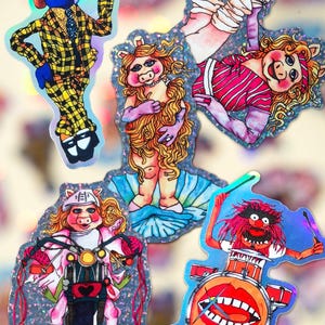 Może przedstawiać: Zestaw sześciu naklejek z postaciami Muppetów, w tym Miss Piggy, Animal, Gonzo, Kermit i dwie wersje Miss Piggy. Naklejki są holograficzne i mają błyszczące tło.