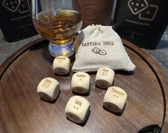 May include: A wooden serving tray holds a glass of amber liquid, tasting dice, and a small burlap bag. The dice are engraved with flavor profiles like "Cinnamon" and "Nutty." The bag reads "TASTING DICE."