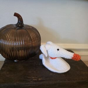 May include: A brown glass pumpkin with a stem sits next to a white crocheted dog with an orange nose and collar. The dog has black eyes and floppy ears. The items are on a dark wooden surface.