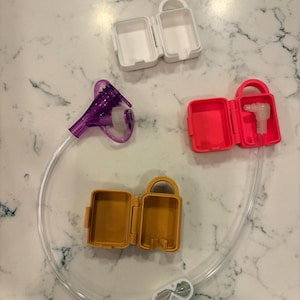 May include: An assortment of colorful plastic medical accessories, including three small, open cases in white, hot pink, and gold, connected by clear tubing. A purple medical device and a white clip are also visible.