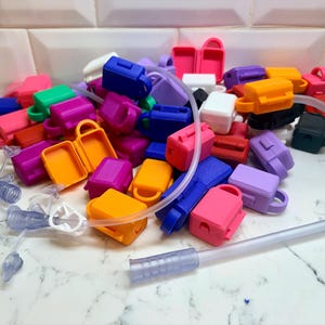 May include: A collection of small, rectangular, multi-colored silicone protective cases for medical tubing connectors. The cases are available in vibrant shades of orange, pink, purple, blue, green, white, and black, featuring attached loops and hinged lids.