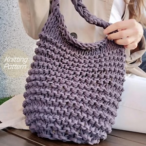 Puede incluir: Un bolso tote tejido a mano, de color lavanda, con un asa trenzada. El bolso presenta un patrón de puntada texturizada y las palabras "Knitting Pattern" son visibles en el lateral. El bolso está diseñado para transportar objetos.
