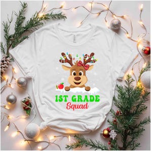 May include: White t-shirt featuring a cartoon reindeer with a red bow, antlers adorned with colorful lights, and an apple. The text "1st Grade Squad" is printed in green and red. Festive design for back-to-school or holiday.