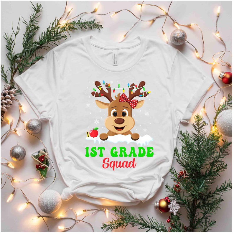 May include: White t-shirt featuring a cartoon reindeer with a red bow, antlers adorned with colorful lights, and an apple. The text "1st Grade Squad" is printed in green and red. Festive design for back-to-school or holiday.
