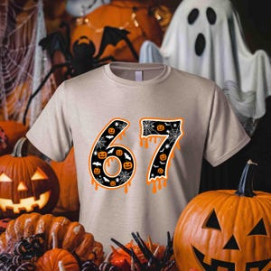 May include: Beige t-shirt with the number "67" in a Halloween-themed design. The numbers are black with orange dripping accents and filled with jack-o'-lanterns and spiderwebs. The shirt is surrounded by pumpkins, spiders, and a ghost.