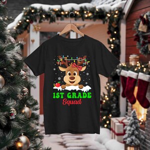 May include: Black t-shirt featuring a cartoon reindeer with a red bow, Christmas lights, and an apple. The text "1st Grade Squad" is printed in green and red. The shirt is hanging on a wooden hanger.