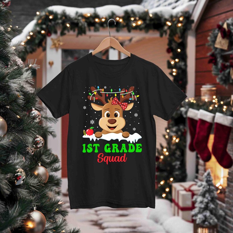 May include: Black t-shirt featuring a cartoon reindeer with a red bow, Christmas lights, and an apple. The text "1st Grade Squad" is printed in green and red. The shirt is hanging on a wooden hanger.