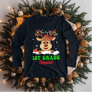 May include: Black long-sleeve shirt with a reindeer graphic. The reindeer has a red bow, Christmas lights, and the text "1st Grade Squad" in green and red. An apple is also included in the design.