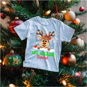 May include: White t-shirt with a Christmas reindeer graphic. The reindeer has antlers decorated with Christmas lights and a red bow. The text "1st Grade Squad" is printed in green and red below the reindeer.