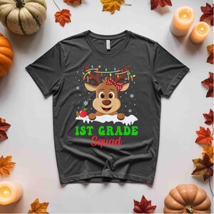 May include: A dark gray t-shirt featuring a cartoon reindeer with antlers adorned with Christmas lights and a red bow. The text "1ST GRADE Squad" is printed in green and red below the reindeer.