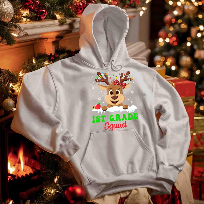 May include: A white hooded sweatshirt featuring a reindeer graphic with antlers adorned with Christmas lights and a red bow. The text "1st Grade Squad" is printed below the reindeer. The sweatshirt has a front pocket.