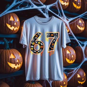 May include: A light gray t-shirt with the number "67" in a Halloween-themed design. The numbers are black with orange and yellow dripping accents, featuring pumpkins, bats, and spiderwebs. The background includes carved pumpkins.