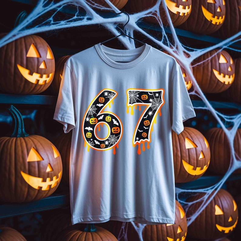 May include: A light gray t-shirt with the number "67" in a Halloween-themed design. The numbers are black with orange and yellow dripping accents, featuring pumpkins, bats, and spiderwebs. The background includes carved pumpkins.