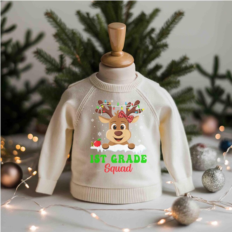 May include: Cream-colored children's sweater featuring a reindeer graphic with a red bow, string lights, and the words "1st Grade Squad" in green. The sweater is displayed on a mannequin with a Christmas theme.
