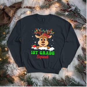 May include: Navy blue sweatshirt featuring a cartoon reindeer with a red bow and string lights on its antlers. The text "1st Grade Squad" is printed below the reindeer in green and red. Snowflakes and an apple are also included in the design.