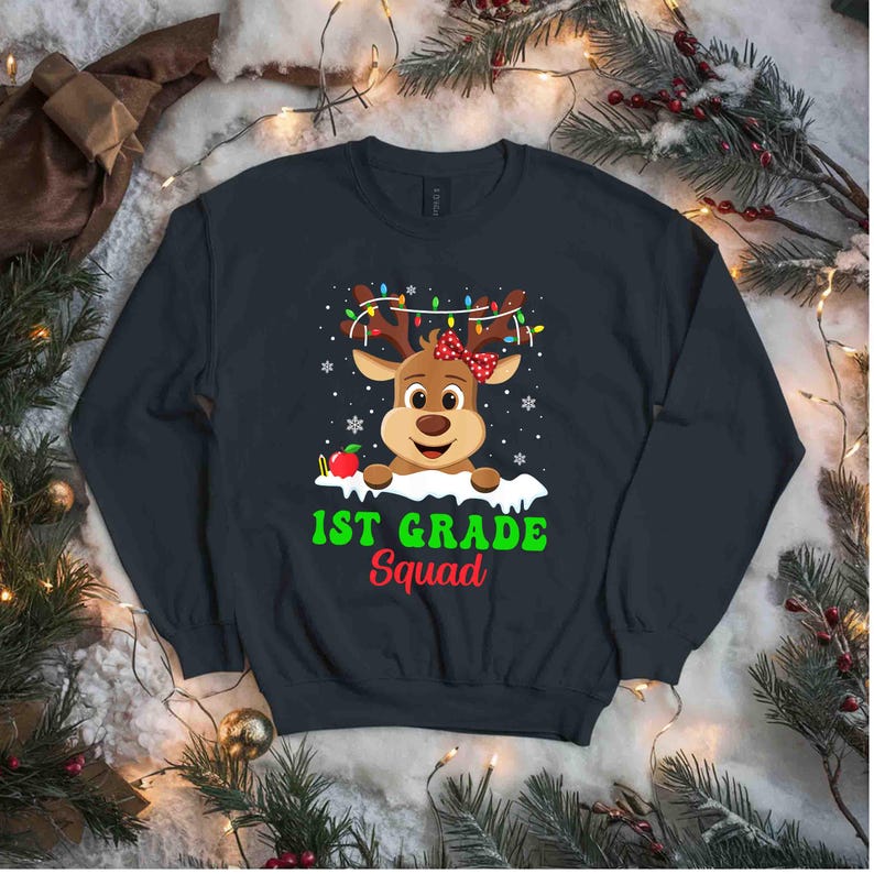 May include: Navy blue sweatshirt featuring a cartoon reindeer with a red bow and string lights on its antlers. The text "1st Grade Squad" is printed below the reindeer in green and red. Snowflakes and an apple are also included in the design.