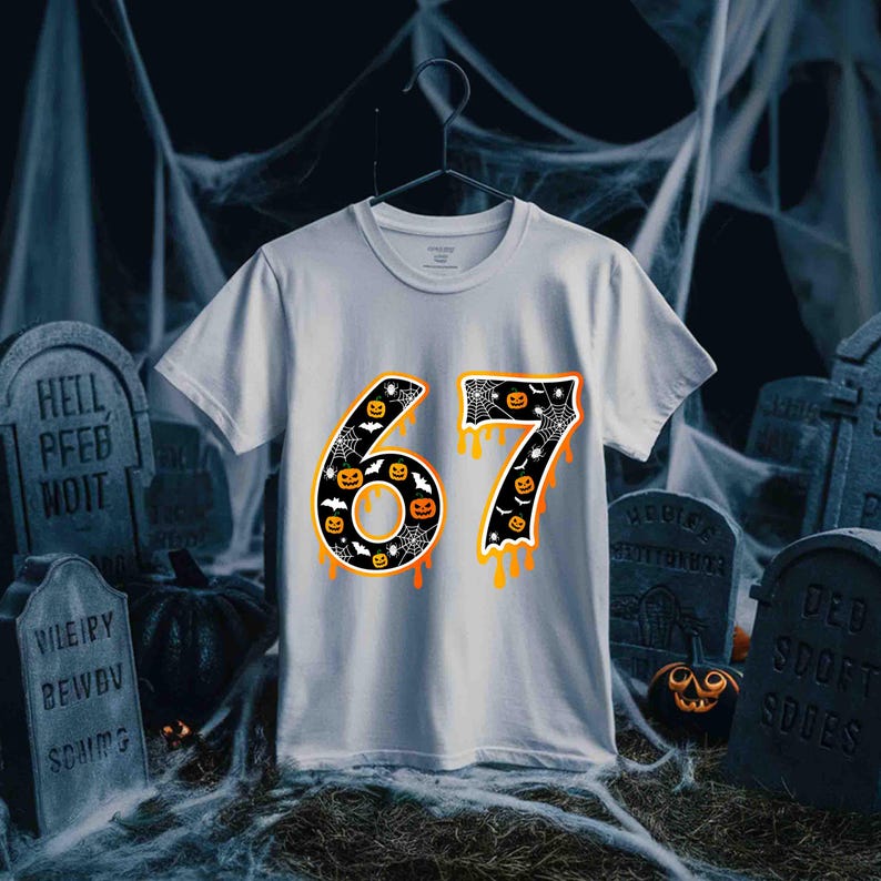 May include: White t-shirt with a Halloween-themed design. The number "67" is prominently displayed, featuring a black background with pumpkins, bats, and spiderwebs. Orange dripping accents add a spooky touch. The shirt is hanging on a black hanger.