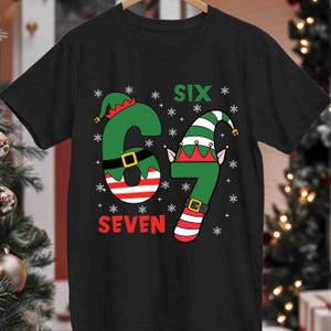 May include: Black t-shirt with a festive design featuring the numbers "6" and "7" styled as elf figures. The word "SIX" is above the "6" and "SEVEN" is below the "7". Snowflakes are scattered around the numbers.