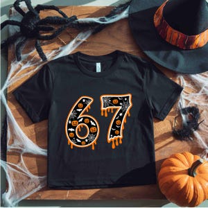 May include: Black t-shirt with the number 67 in a Halloween-themed design. The numbers are outlined in orange with a dripping effect, filled with spiderwebs and jack-o'-lanterns. A witch's hat and pumpkin are in the background.