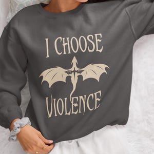 Woman wearing a comfy white, unisex crewneck sweatshirt. Striking graphic design on the front features a majestic Black Dragon, boldly proclaiming the phrase I Chose Violence, a statement that's sure to resonate with fans of intense, passionate relationships.