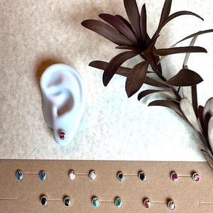 May include: A display of 16 small, oval-shaped earrings with various gemstone colors, including red, blue, green, and white. The earrings are arranged in two rows on a brown surface. A white plaster ear model is shown with a red gemstone earring.