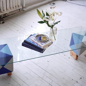May include: A glass coffee table with a clear top and a geometric patterned base. The base is made of blue, white, and gray painted wood. The table is decorated with a vase of white lilies and a stack of books.