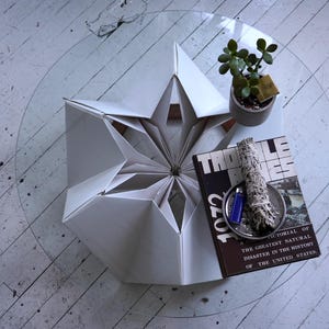 May include: A white origami-style table with a starburst design. A book titled "TROLE" is on the table, along with a small potted plant and a sage bundle.