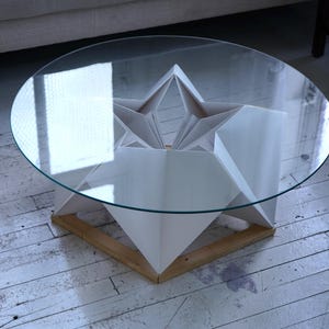 Op de afbeelding: Een witte origami-achtige salontafel met een helder glazen blad. De tafel heeft een stervormig ontwerp en staat op een houten onderstel.