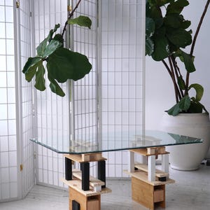 Puede incluir: Una mesa con tapa de vidrio y una base de madera única de varios niveles. La base está hecha de madera natural con un acabado marrón claro y presenta detalles en blanco y negro. La mesa está sobre un suelo blanco.