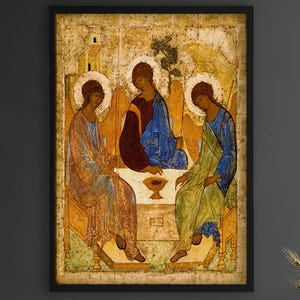 May include: A framed wall art print featuring a reproduction of the Trinity icon. The artwork displays three figures seated around a table with a central chalice, rendered in gold, blue, and red tones with a weathered, aged texture and black frame.