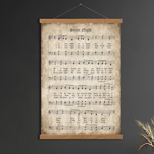 May include: A vintage-style wall hanging featuring the sheet music for "Silent Night." The text is printed on aged paper, and the wooden bars at the top and bottom add a rustic touch. The artwork is suspended by a simple string.