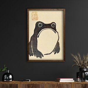 May include: Framed print featuring a stylized black and brown frog illustration. The artwork has a minimalist aesthetic with a cream-colored background and a brown wooden frame. A red and gold stamp is in the upper right corner.