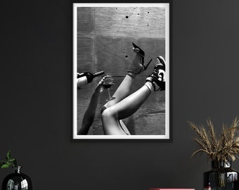 Op de afbeelding: Zwart-wit ingelijste print met een persoon die wijn in een glas schenkt. De afbeelding toont benen met hoge hakken en sneakers. De print wordt weergegeven op een donkere muur boven een houten kast.