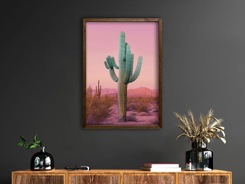 Może przedstawiać: Oprawiony wydruk przedstawiający duży kaktus saguaro na pustynnym krajobrazie. Kaktus ma turkusowy kolor na tle r&oacute;żowo-fioletowego nieba. Grafika jest wyświetlana na ciemnoszarej ścianie.