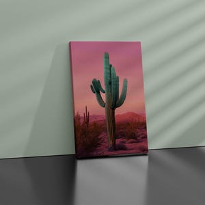 Może przedstawiać: Wydruk na pł&oacute;tnie przedstawiający pustynny krajobraz z dużym kaktusem saguaro pod r&oacute;żowo-pomarańczowym zachodem słońca. Kaktus jest zielony, a tło ukazuje g&oacute;ry i pustynną roślinność.