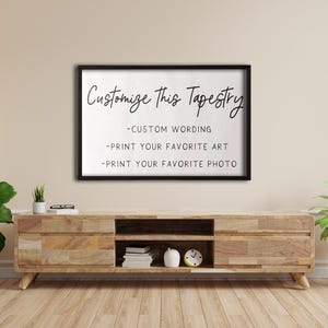 Op de afbeelding: Een zwart ingelijst bord met witte tekst die "Pas deze wandtapijt aan - Aangepaste tekst - Print je favoriete kunst - Print je favoriete foto" luidt. Het bord hangt aan een beige muur boven een houten plank met een klok en boeken.