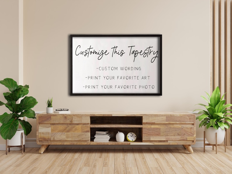Op de afbeelding: Een zwart ingelijst bord met witte tekst die "Pas deze wandtapijt aan - Aangepaste tekst - Print je favoriete kunst - Print je favoriete foto" luidt. Het bord hangt aan een beige muur boven een houten plank met een klok en boeken.