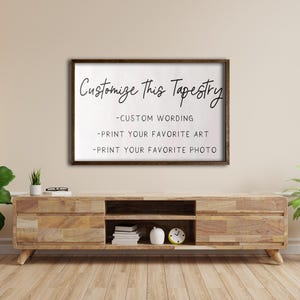 Op de afbeelding: Een bruin houten frame met een witte achtergrond en zwarte tekst die "Customize This Tapestry - Custom Wording - Print Your Favorite Art - Print Your Favorite Photo" luidt. Het frame hangt aan een beige muur boven een houten entertainmentcenter.