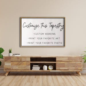 Op de afbeelding: Een wit ingelijst bord met de tekst "Customize This Tapestry" en drie opsommingstekens: "Custom Wording", "Print Your Favorite Art" en "Print Your Favorite Photo". Het bord hangt aan een lichtbeige muur boven een houten plank.