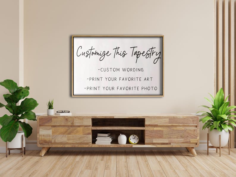 Op de afbeelding: Een wit ingelijst bord met de tekst "Customize This Tapestry" en drie opsommingstekens: "Custom Wording", "Print Your Favorite Art" en "Print Your Favorite Photo". Het bord hangt aan een lichtbeige muur boven een houten plank.
