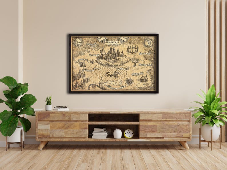K&ouml;nnte beinhalten: Eine gerahmte Karte von Hogwarts mit detaillierten Illustrationen des Schlosses und seiner Umgebung. Die sepiafarbene Karte in einem schwarzen Rahmen wird an einer hellen Wand &uuml;ber einem Holzschrank ausgestellt.
