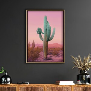 Może przedstawiać: Oprawiony wydruk przedstawiający kaktus saguaro na pustynnym krajobrazie. Kaktus ma żywy turkusowy kolor, osadzony na r&oacute;żowo-fioletowym niebie o zachodzie słońca. Grafika jest wyświetlana na ciemnoszarej ścianie.